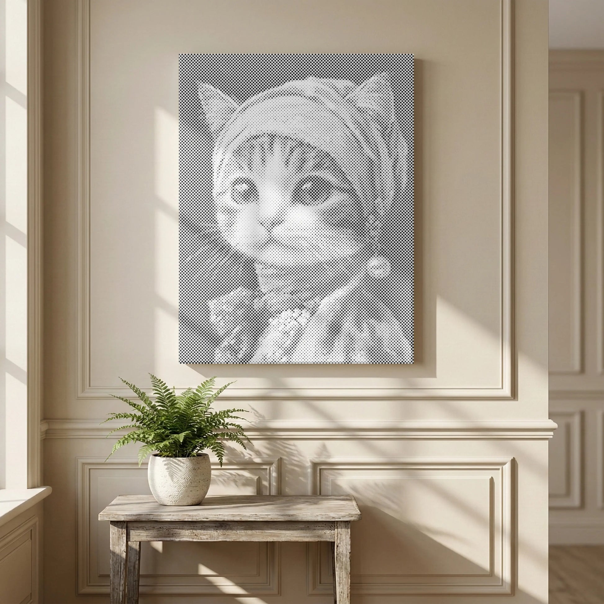 A vertical canvas wall art print featuring a black and white portrait of a kitten wearing a headscarf and pearl earring, displayed on a beige paneled wall above a small wooden table with a potted fern.
