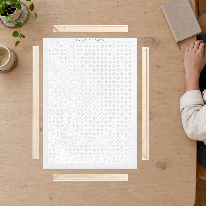 Kit cadre en bois pour toile, sur une table de loisirs créatifs, avec la main d’une personne posée au bord.